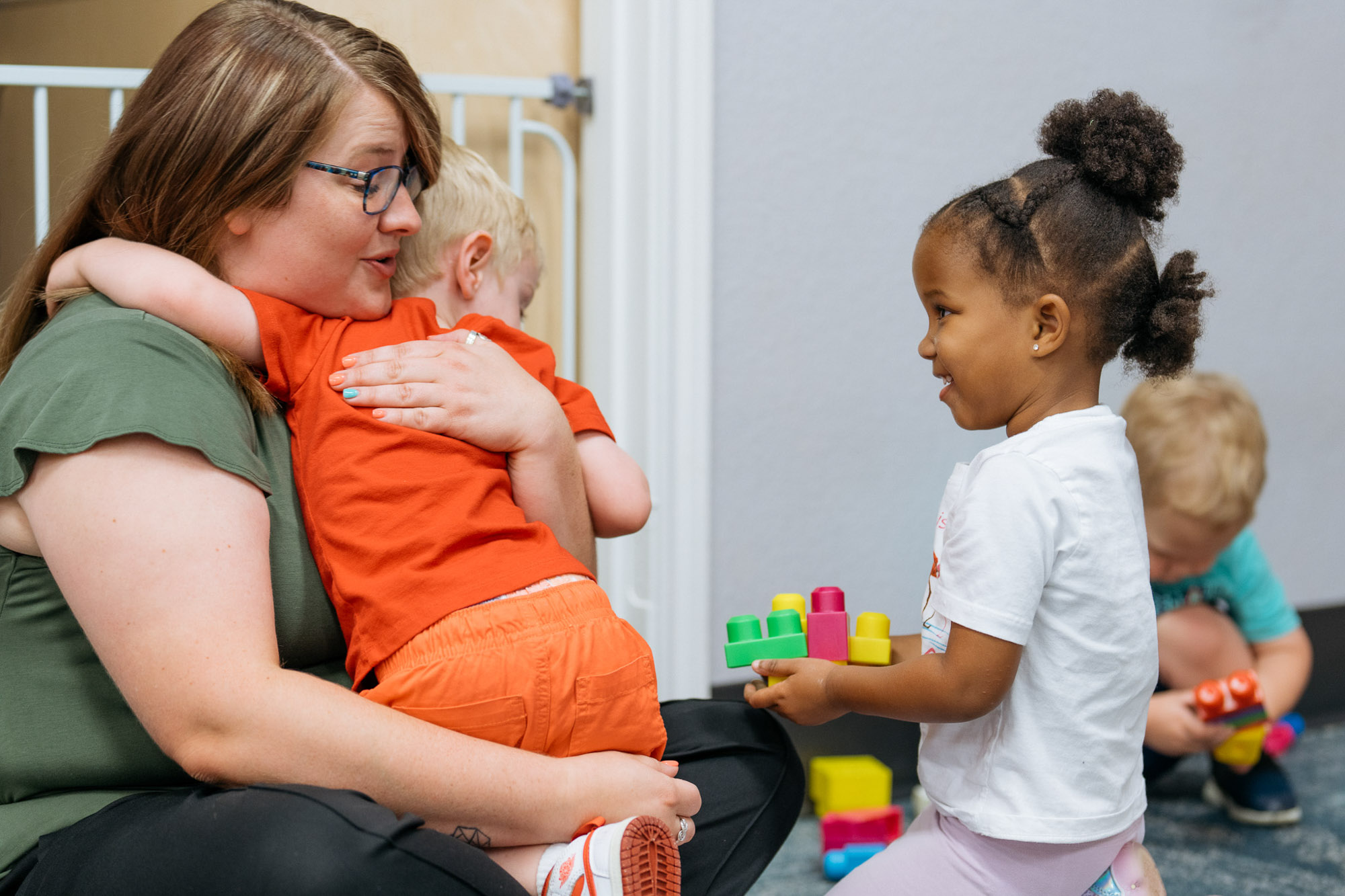 An adult hugs a child in an orange outfit while another child stands nearby holding colorful plastic blocks. A third child in the background also plays with toys.