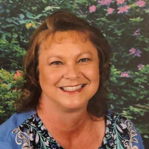 Woman smiling in front of a backdrop with green foliage and purple flowers, wearing a blue top with a colorful embroidered pattern.