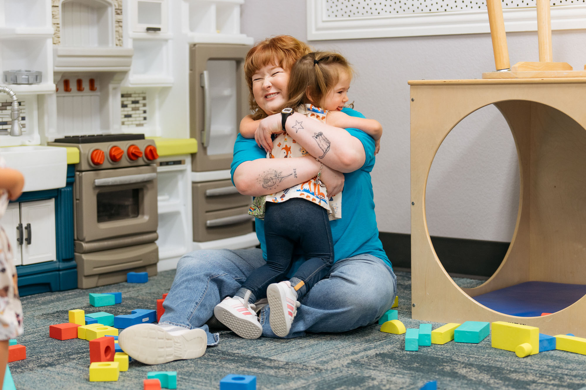 An adult and a child sit on the floor and hug each other in a playroom surrounded by colorful plastic blocks and play kitchen equipment.